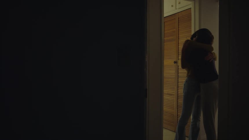 Two women hug each other tightly while standing in a hallway near closet doors