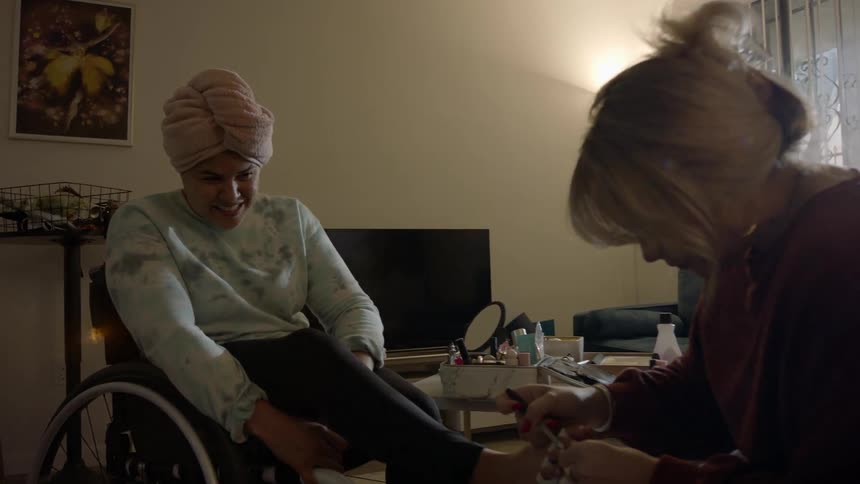 A teenage girl sits in a wheelchair at home and talks as a woman paints her toe nails