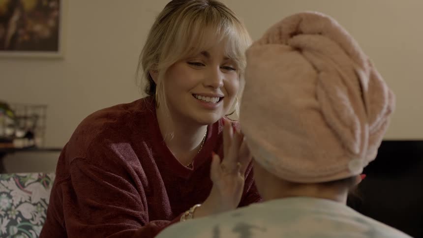 Blonde woman smiles and gestures while assisting woman with skincare