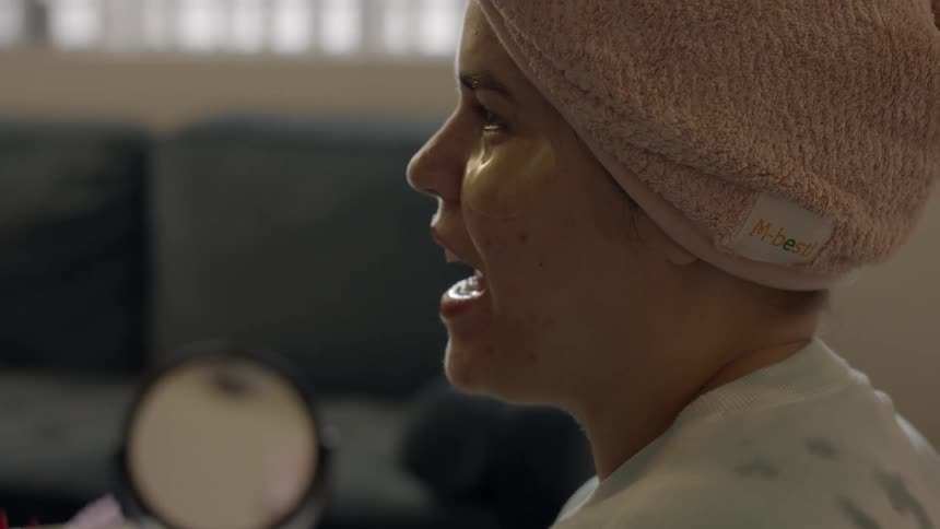 Young woman wearing a towel over her head speaks while a friend applies makeup on her face