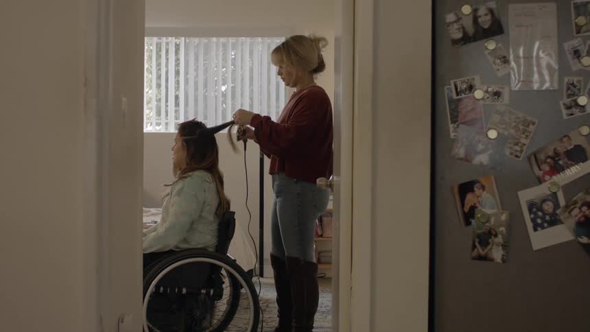 A woman sits in a wheelchair as another woman stands behind her curling her hair