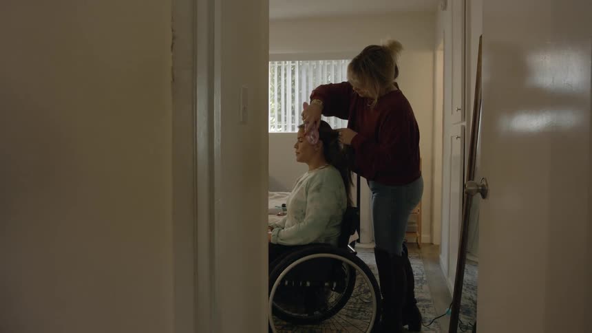 Woman combing the hair of a friend sitting on a wheelchair inside a room
