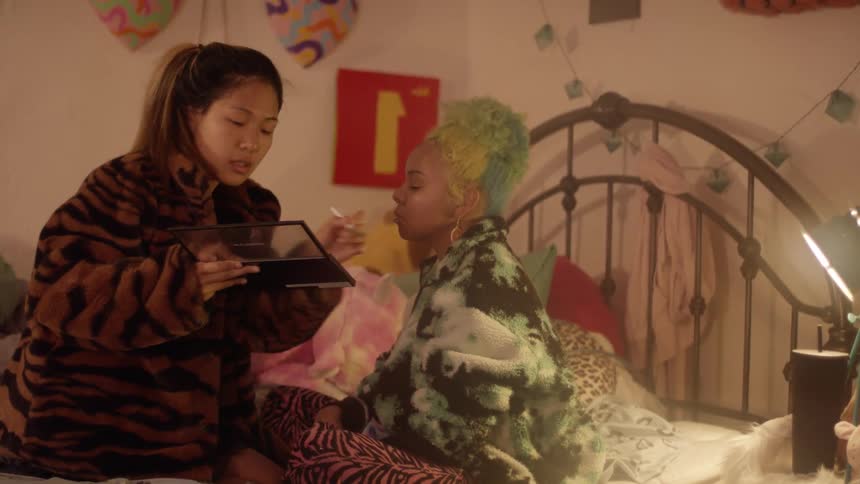 A woman carefully applies eye shadow to another woman while they sit on a bed in a messy and colorful bedroom
