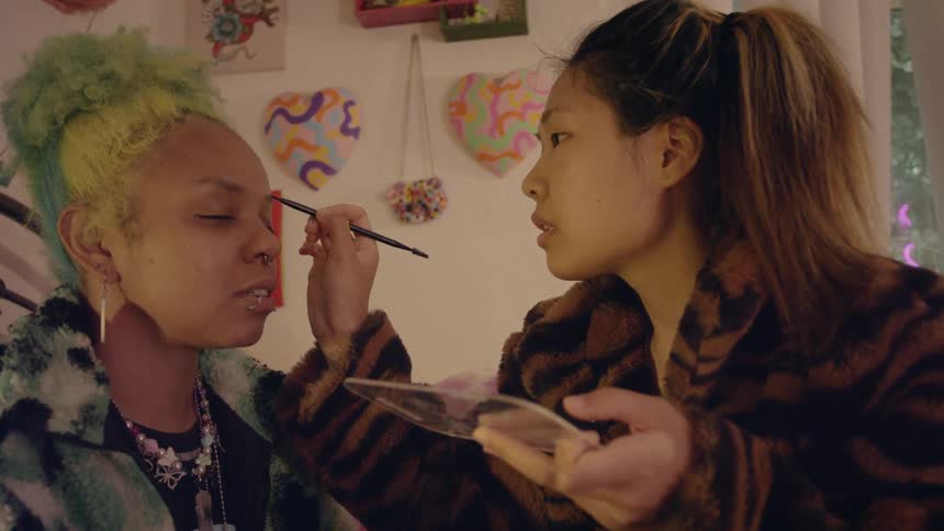 An Asian woman and a black woman are sitting indoors and applying makeup