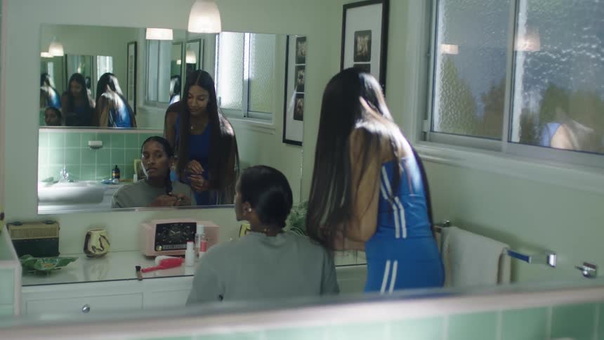 Young woman helping a friend braid her hair in front of an infinite mirror inside the bathroom