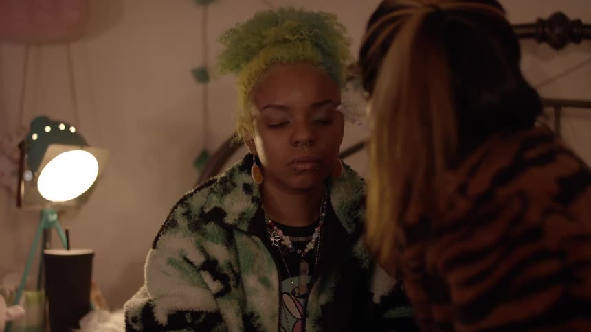 Stylist applying makeup on the eyelids of a woman with green curly hair sitting in a room