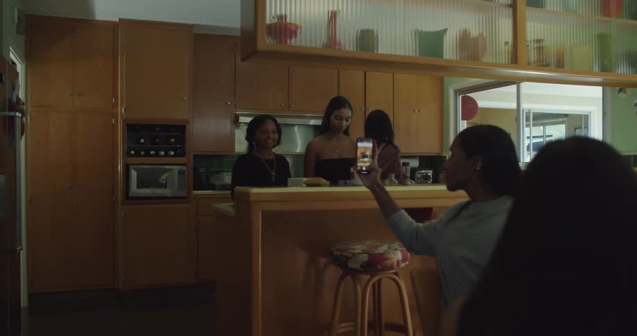 A group of women are having their picture taken in the kitchen by another woman sitting in the next room