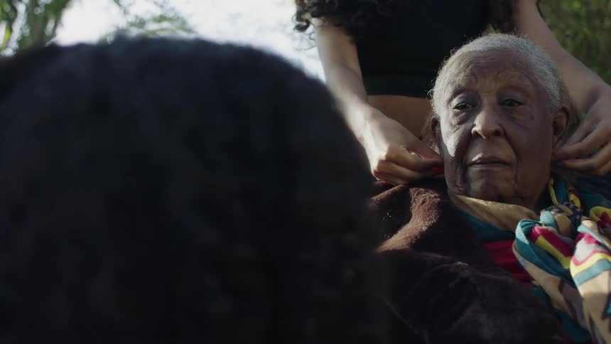 Woman with curly hair black t shirt touches elderly womans gray hair sitting in chair and talking
