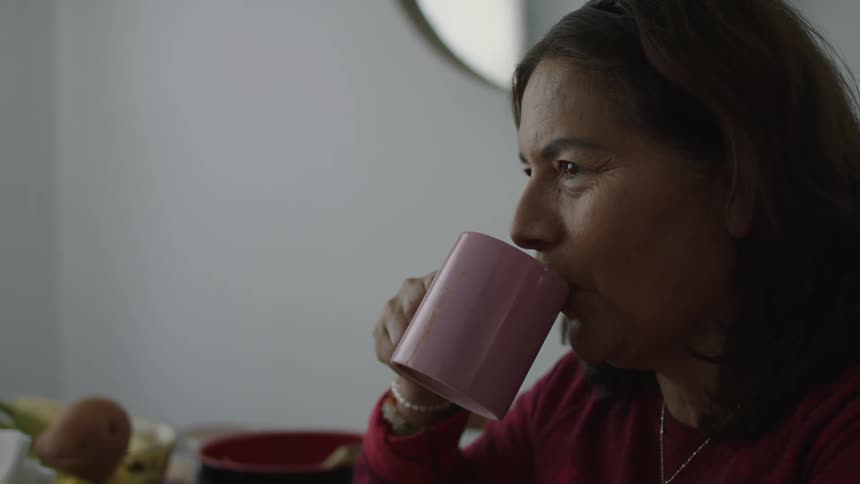 Middle aged woman with wavy hair and red sweater drinks from pink cup talking to person sitting at table