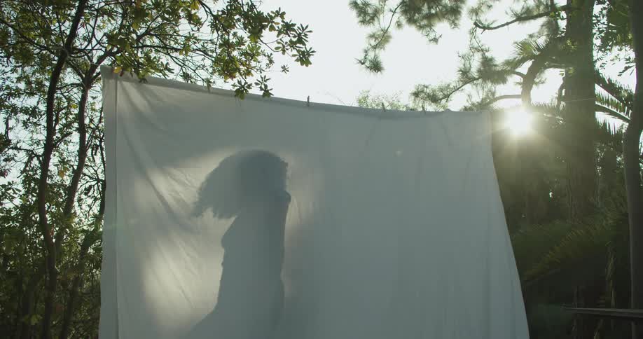 Silhouette shadow of a young girl walking behind a white bed sheet hanging in the garden at morning