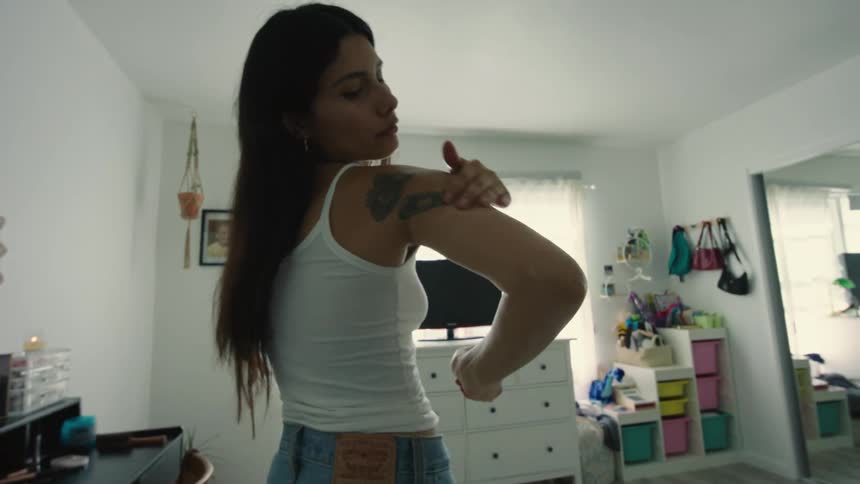 A woman applying lotion to her tattooed shoulder and arm in a bedroom