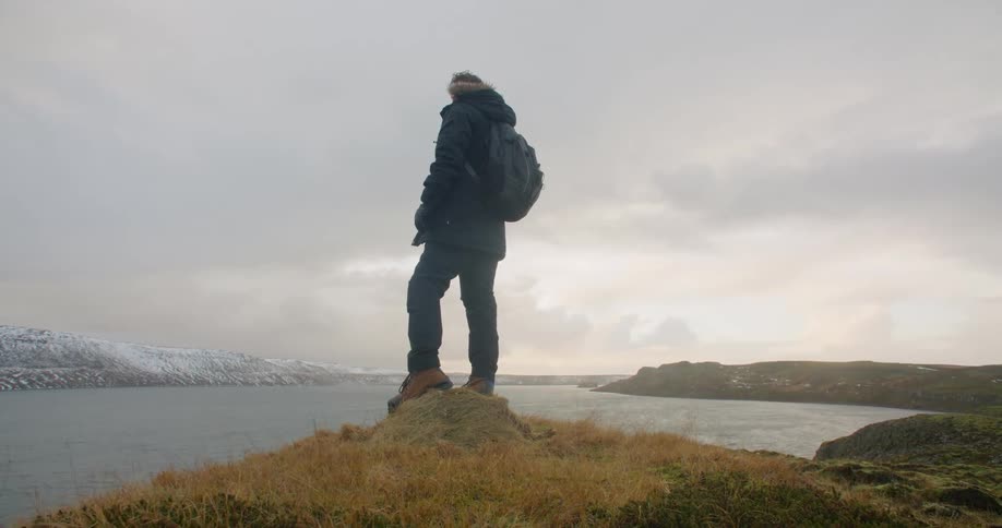 Man wearing coat and carrying backpack stands overlooking water and opposing snow covered shore