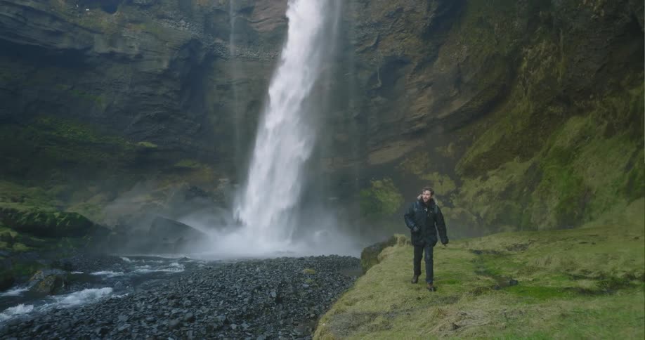 A man is walking through a canyon below a waterfall cascading down rocks