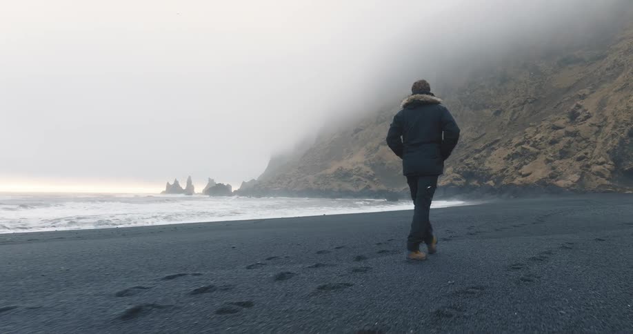 A man in a winter coat walks across a beach with dark soil towards a mountain shrouded in fog