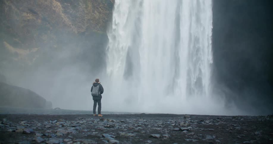 Waterfalls flow down the side of a cliff as a man walks nearby and stops to look at the water