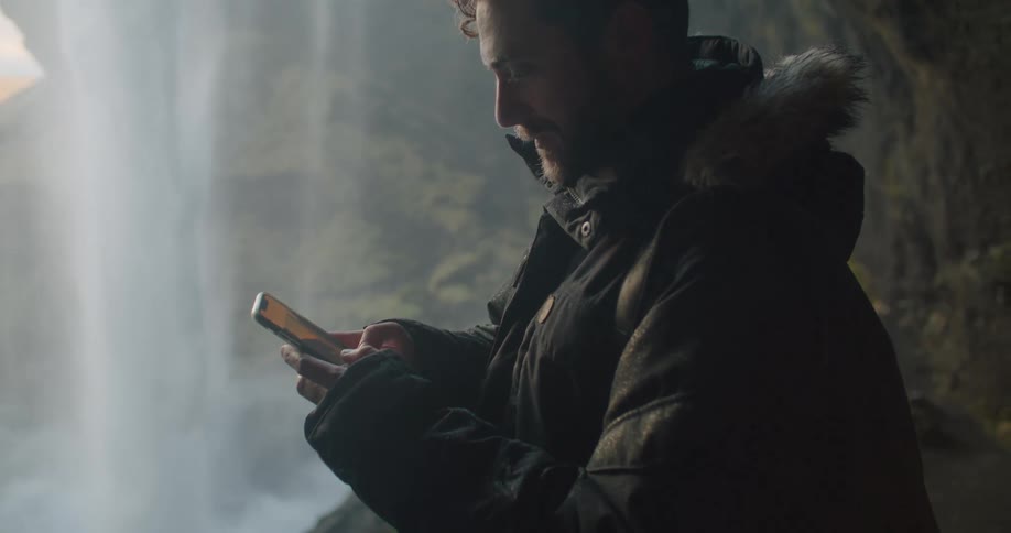 Young man takes a picture of a waterfall from the entrance of a cave in the mountains