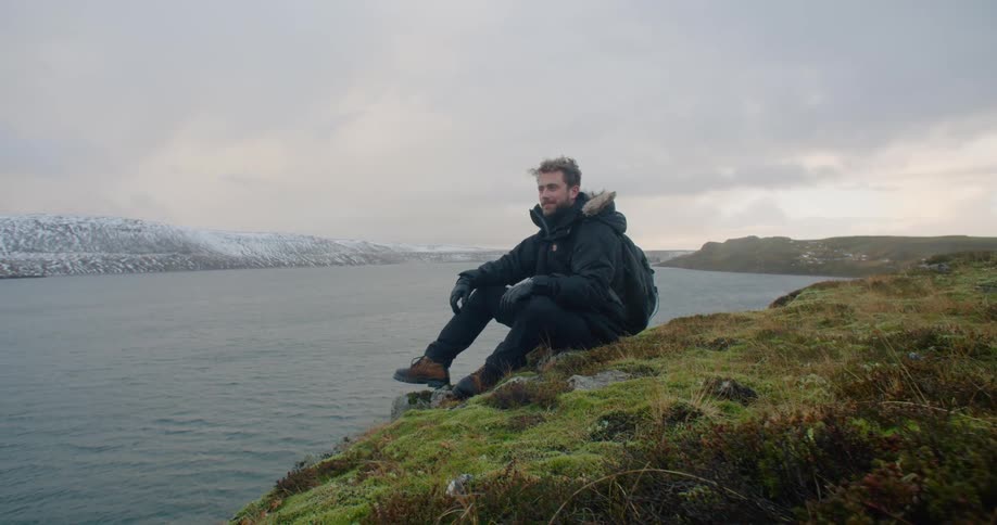 Young bearded man sitting on the coast close to an icelandic fjord at sunset