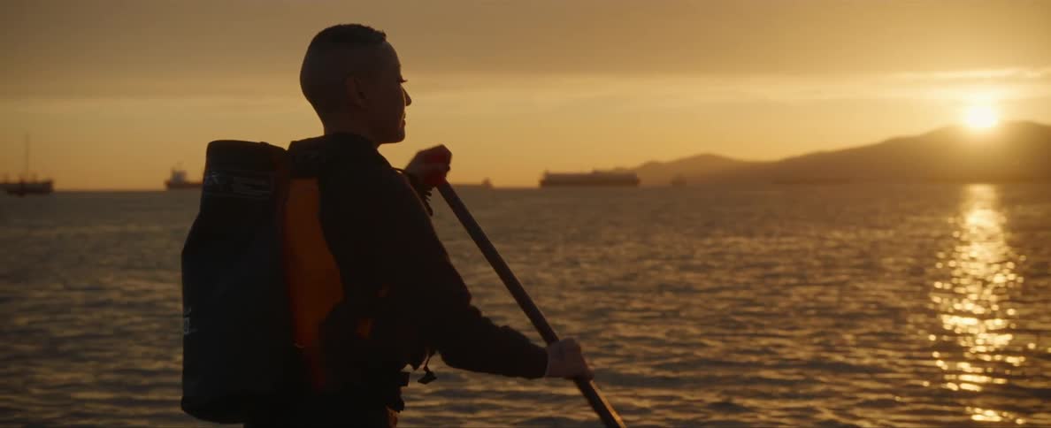 A man is standing in a boat rowing as the sun shines brightly over a mountain and onto the water