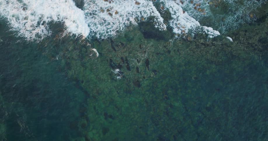 Aerial view of waves rolling across clear water to the coast and the rocks below are visible