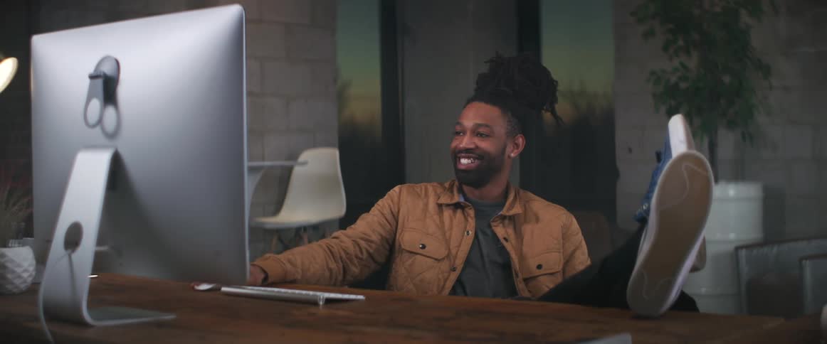 A man laughs while using his computer with his legs propped on his desk