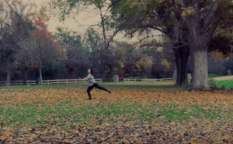 Woman with gray shirt and black leggings dances on lawn with brown leaves in public park