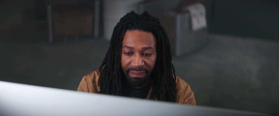 A man sitting at a computer in an office smiles and grins at the monitor