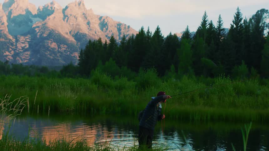 A lone man fishing in a river near mountains on a summer morning