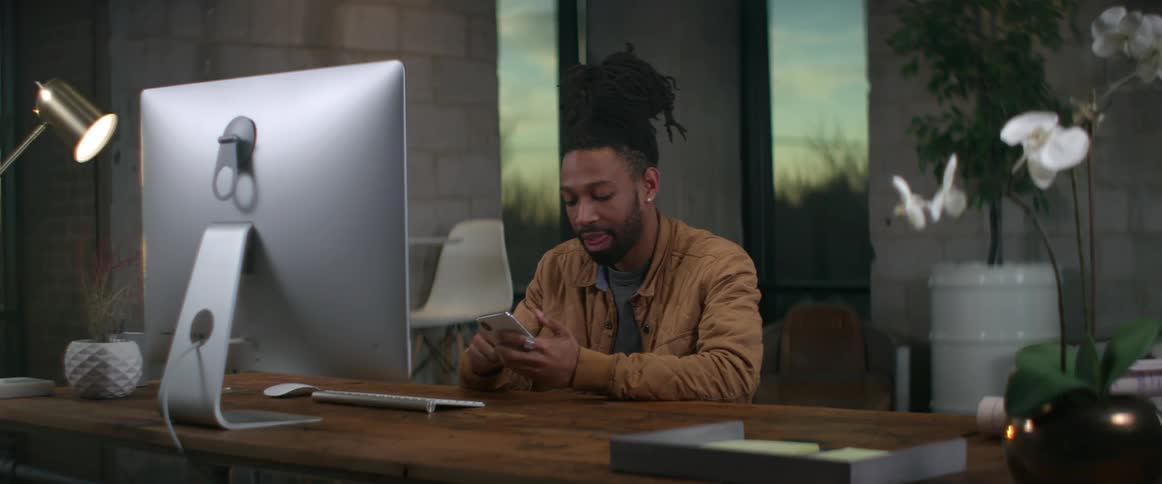 A man sits at his desk in front of a computer and smiles and laughs as he uses his cell phone