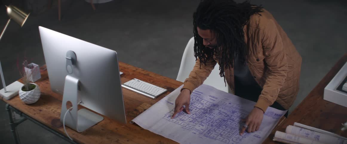 Man leans over table examining blueprint set on surface near computer