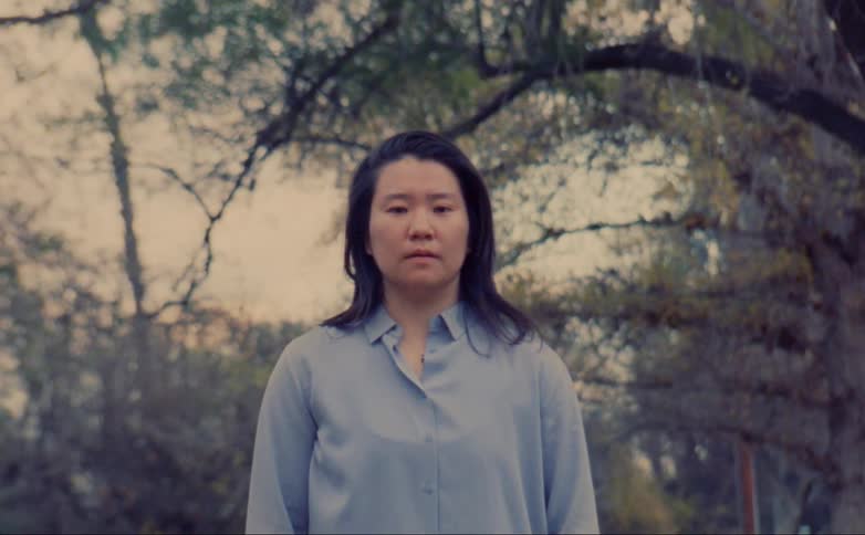 Woman stands still watching at the camera near the trees on the park close to the highway