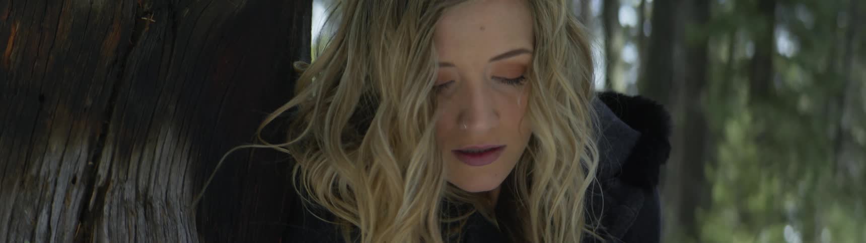 A teenage girl looks down while leaning against a tree in a forest and then slowly turns her head and look up with a worried expression