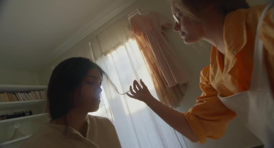 A girl twirls her friends hair in a bedroom