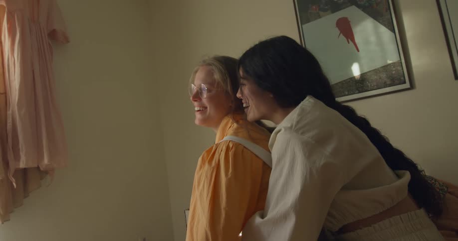 Woman with white shirt holds woman wearing glasses and orange shirt sitting on bed talking and laughing in room