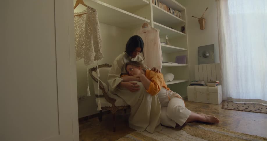 Woman strokes head of second woman sitting on the floor and cuddling on her lap