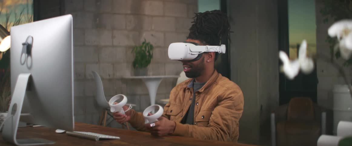 A man sits in front of his computer at home and plays a virtual reality game with a headset on