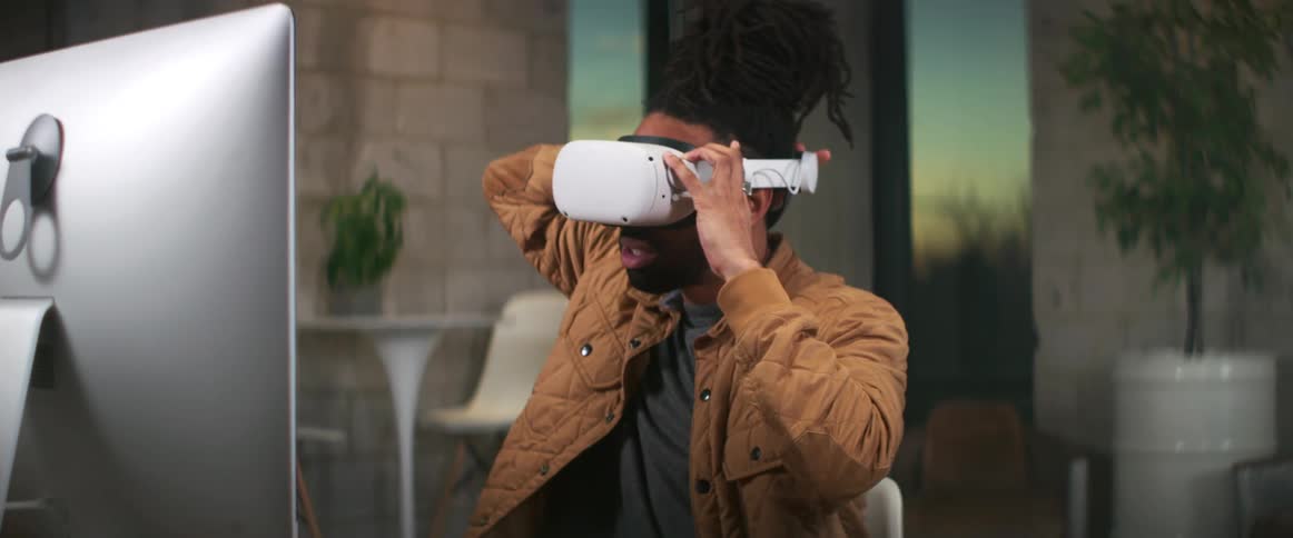 Man with braids brown jacket wears vr headset sitting at desktop looking at computer screen