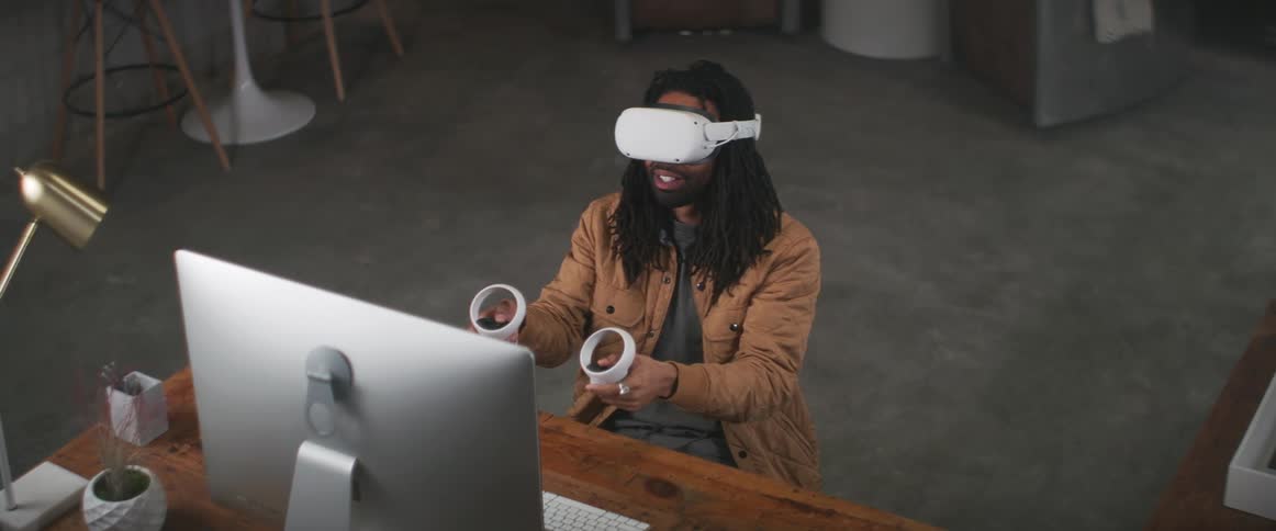A black man sitting at a desk and he pulls a virtual reality headset down over his eyes and holds the game controllers in his hands