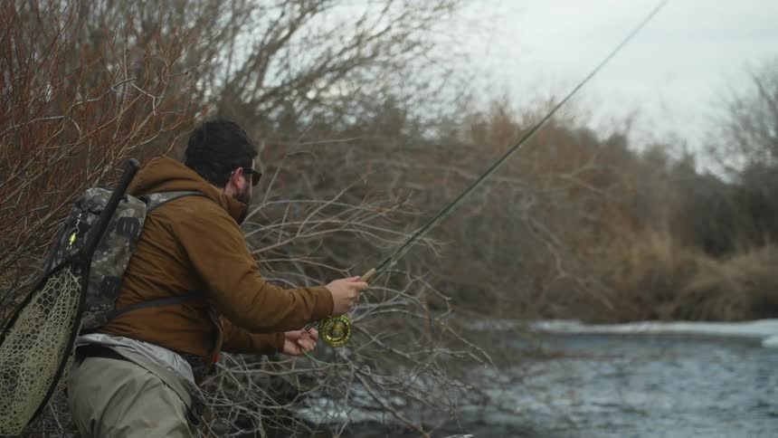 A fisherman is standing on a river bank near bare trees and casting his line
