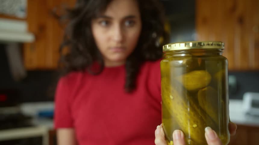 Young woman intensely looking at a pickle jar she is holding on her hands in the kitchen
