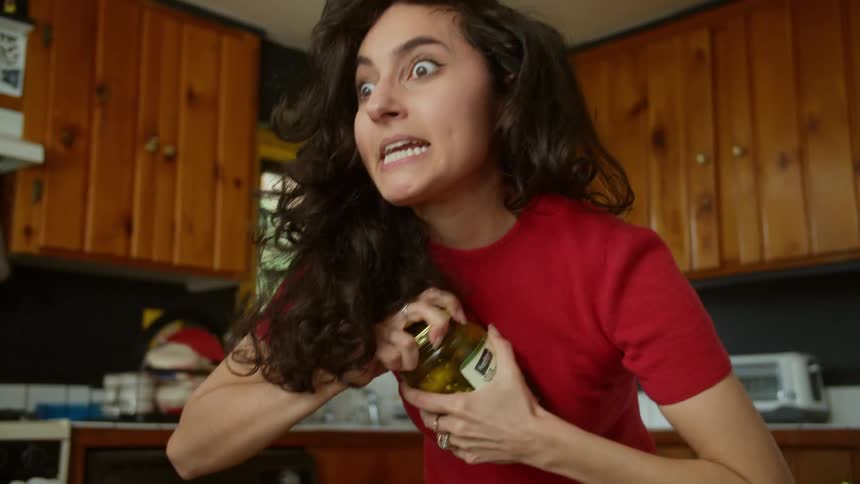 Woman standing in kitchen yells as she strains to open pickle jar