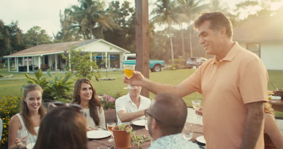 A man standing at the outdoor dining table leading the family group in a toast