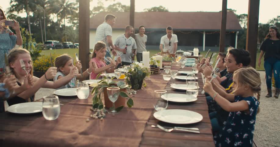 A group of children sitting at an outdoor dining table banging their cutlery and chanting while their parents look on