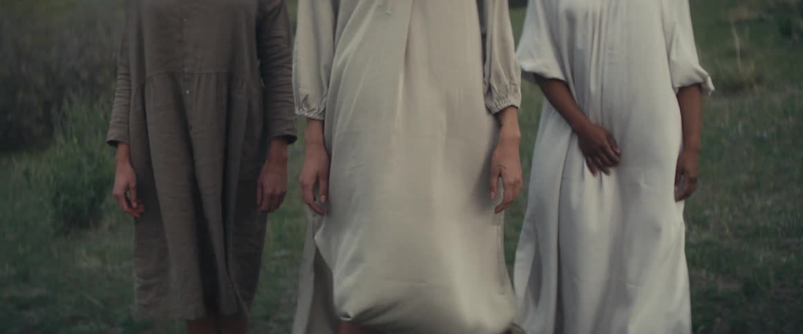Three women wearing dresses stand on sloping grassy field