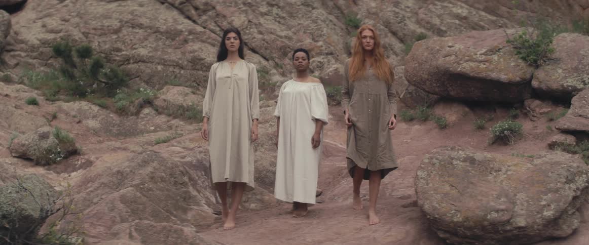 Three women with white gray and brown dresses walks on rocky hill posing for camera