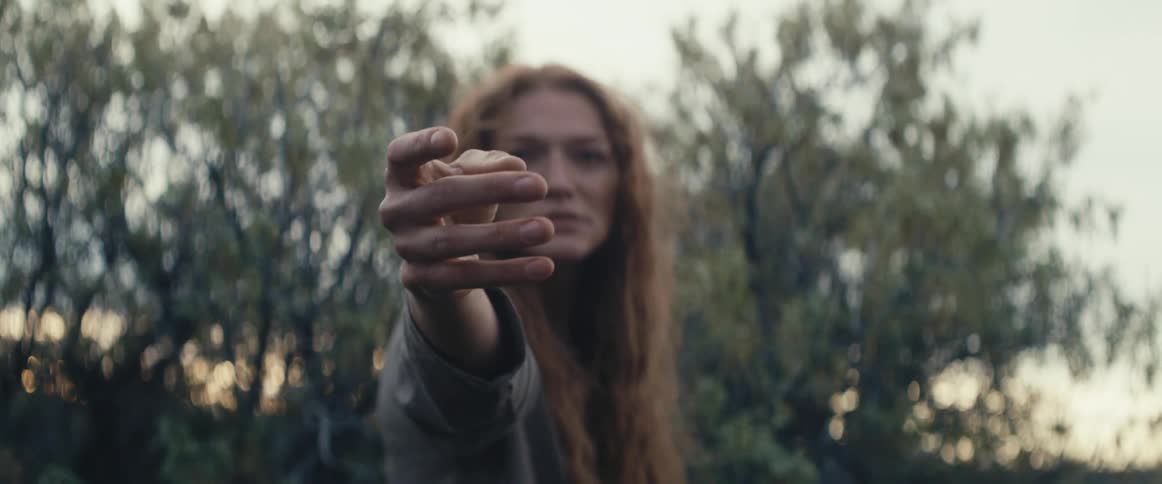 A woman in a wooded area stands and stretches her arm out in front of her