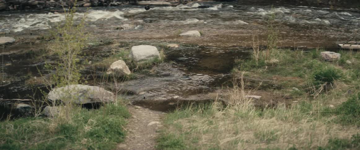 A grass covered bank is on the side of a river with water flowing over rocks