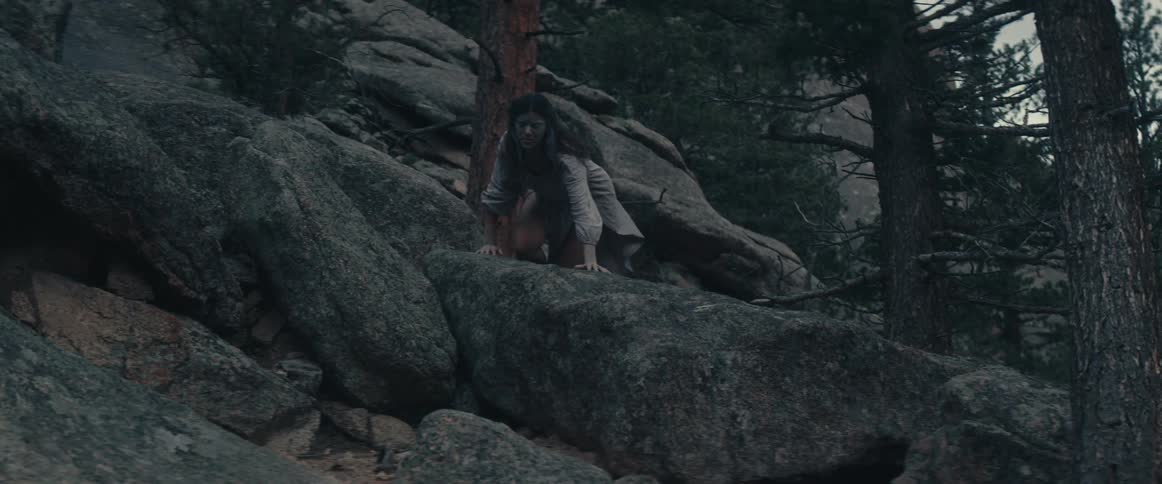 A woman slithers and crawls from behind rocks in a canyon