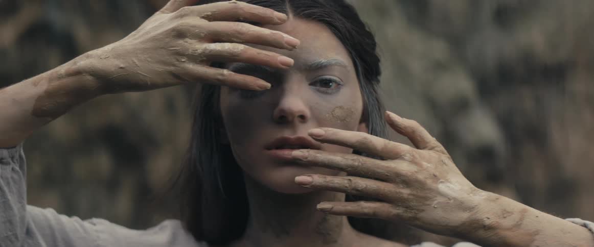 A teenage girl caked in dirt stands in front of a cliff with her hands in front of her face and slowly moves her hands away