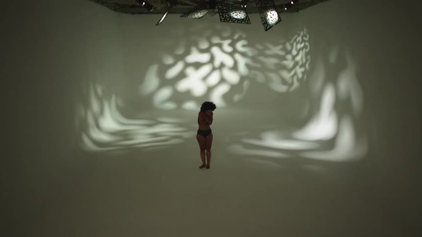 Woman moves and poses on stage set backed by pattern cast from light as spotlight turns on and off