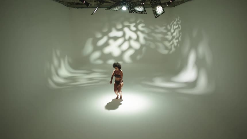 Spotlight highlights person standing in white room in front of light pattern as she squats and stands